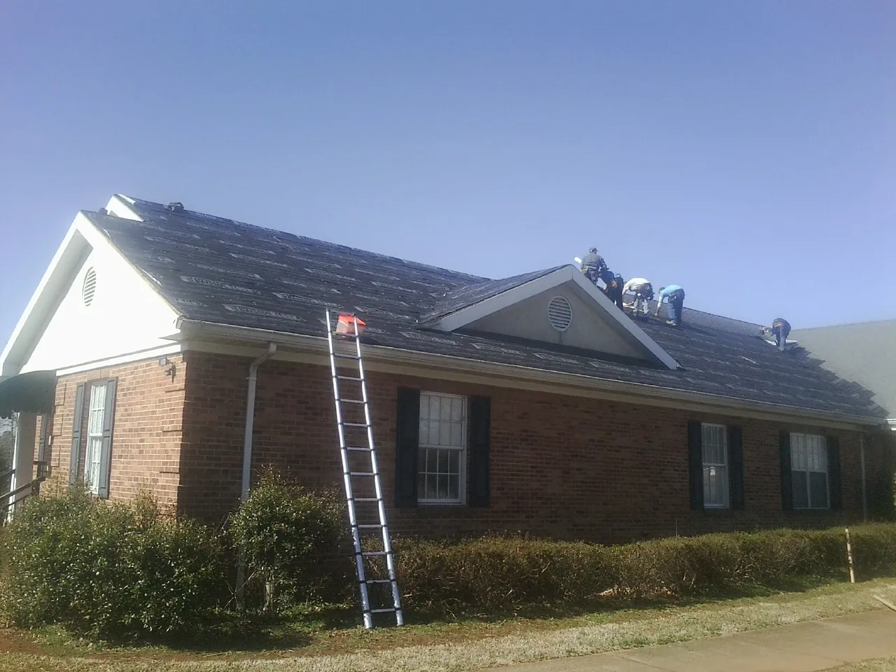 Crew tearing shingles off a brick ranch roof with a ladder set — Emergency Roof Repair work by our crew in Hampton Bays