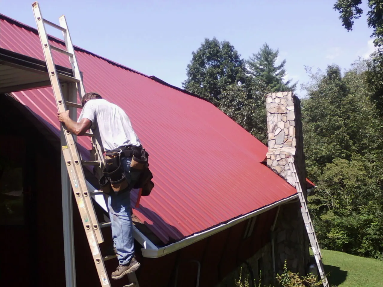 Roofer on a ladder beside a red metal roof and stone chimney — Soffit & Fascia Repair by Roofing Brothers in Hampton Bays