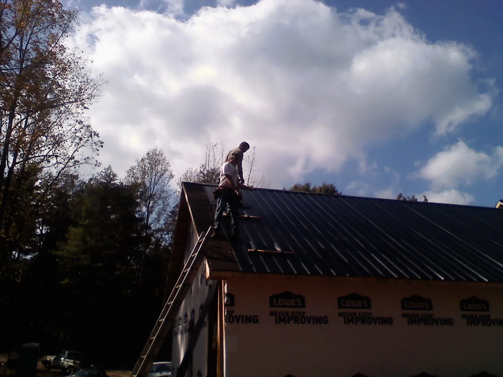 Roofer fastening dark metal panels high against a cloudy sky, from a Roof Inspection job in Hampton Bays